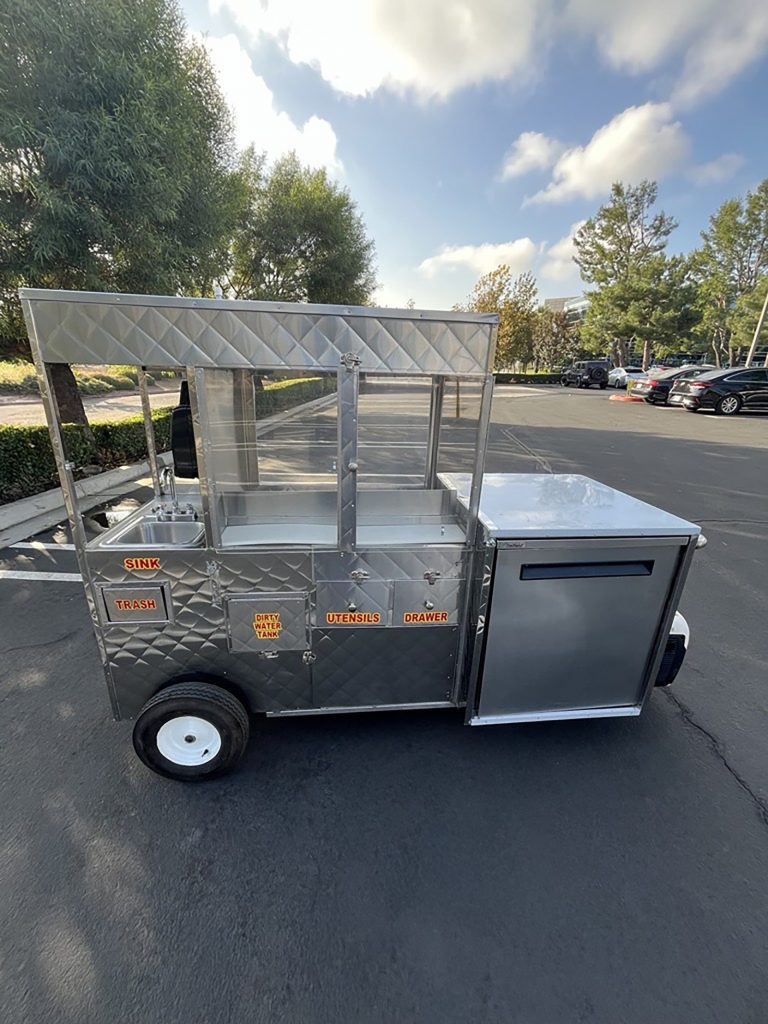 A stainless steel mobile food cart parked outdoors on a paved lot. The cart is labeled with orange text indicating compartments for a sink, trash, dirty water tank, utensils, and a drawer.